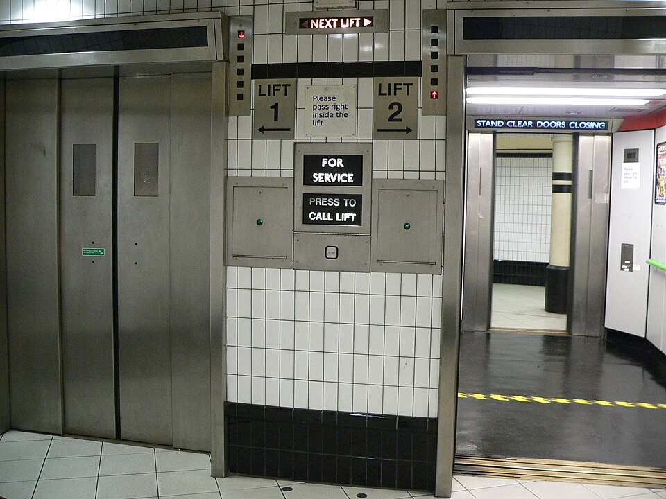 Two elevators in the lower level of a London Underground station. The arrows show each elevator's position and direction of travel. The elevator on the right is preparing to ascend, and the left elevator is descending from the top floor.