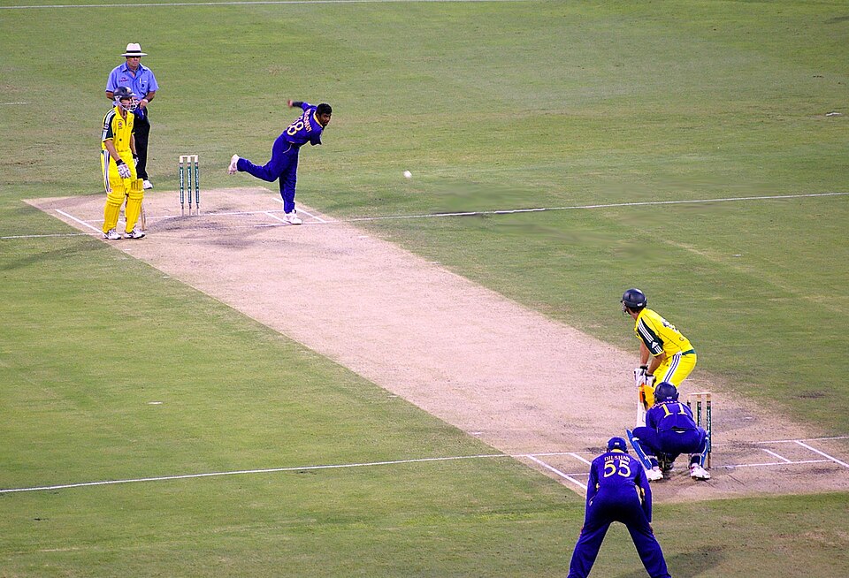 Muttiah Muralitharan (Murali) of Sri Lanka bowls to Adam Gilchrist of Australia in a Twenty20 International match. The pale area of the field is the pitch, with a wicket in place at each end. The batter (Gilchrist) is using his bat to defend the wicket against the white ball which Murali has just bowled. Sri Lanka's wicket-keeper (#11) is crouching just behind the wicket which Gilchrist is defending. The other Sri Lanka player (#55) is a fielder operating in the "first slip" position. Gilchrist's partner, the "non-striker", stands by the other wicket at the bowler's end of the pitch. Behind that wicket stands one of the two umpires. The lines on the pitch are the various creases.